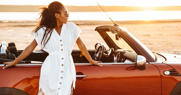 Woman lounging against rust-colored convertible parked overlooking lake or ocean. Used as featured image on Sixt car rental coupons, deals, and offers.