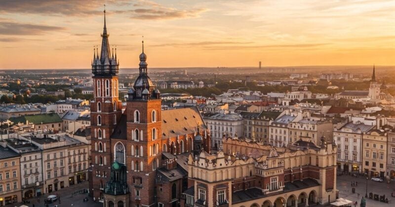 Aerial view of Krakow, Poland, bathed in the warm, golden light of a setting or rising sun. The central focus is the iconic St. Mary's Basilica, with its two differently sized towers, standing majestically in the heart of the city's Main Market Square (Rynek Główny).