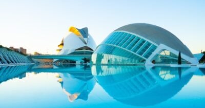 A modern architectural complex, the City of Arts and Sciences in Valencia, Spain, features futuristic white and glass buildings, including the Hemisfèric, which is shaped like an eye. The structures are beautifully reflected in a serene, blue-toned pool of water under a clear, bright sky.