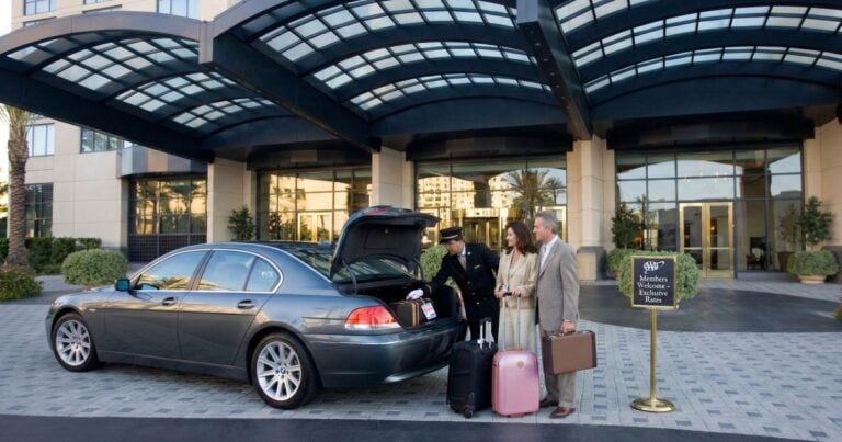 A valet unloads luggage from the trunk of a dark grey BMW sedan for an older couple at the grand entrance of a luxury hotel. A brass sign with the AAA logo next to them reads "Members Welcome - Exclusive Rates."