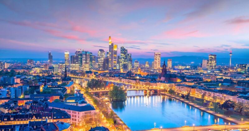 Aerial twilight view of the Frankfurt, Germany skyline and the Main River with glowing city lights and a purple sunset sky.