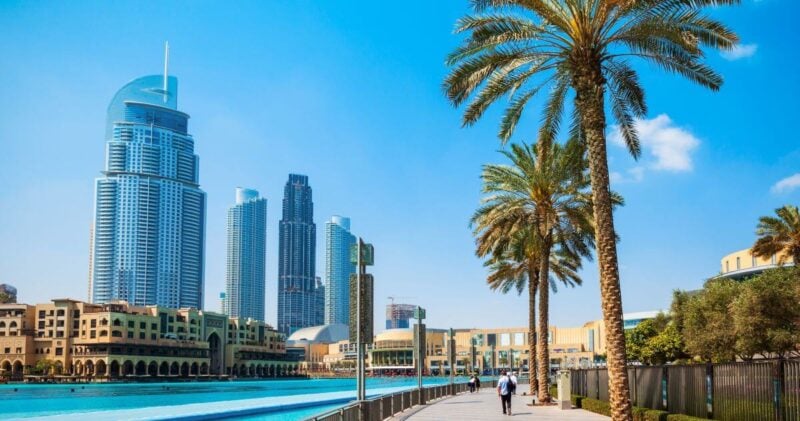 Sunny promenade in Downtown Dubai lined with palm trees, overlooking the Burj Khalifa lake with modern skyscrapers in the background.