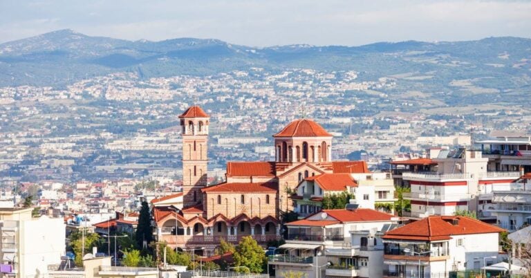 Aerial panoramic view of Thessaloniki, Greece, featuring the historic White Tower on the waterfront and the dense city landscape under a bright sky, overlooking the blue waters of the Thermaic Gulf.