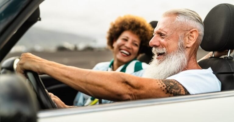 Senior couple laughing while on the road in a convertible. Featured image for How to Get Senior Car Rental Discounts article.